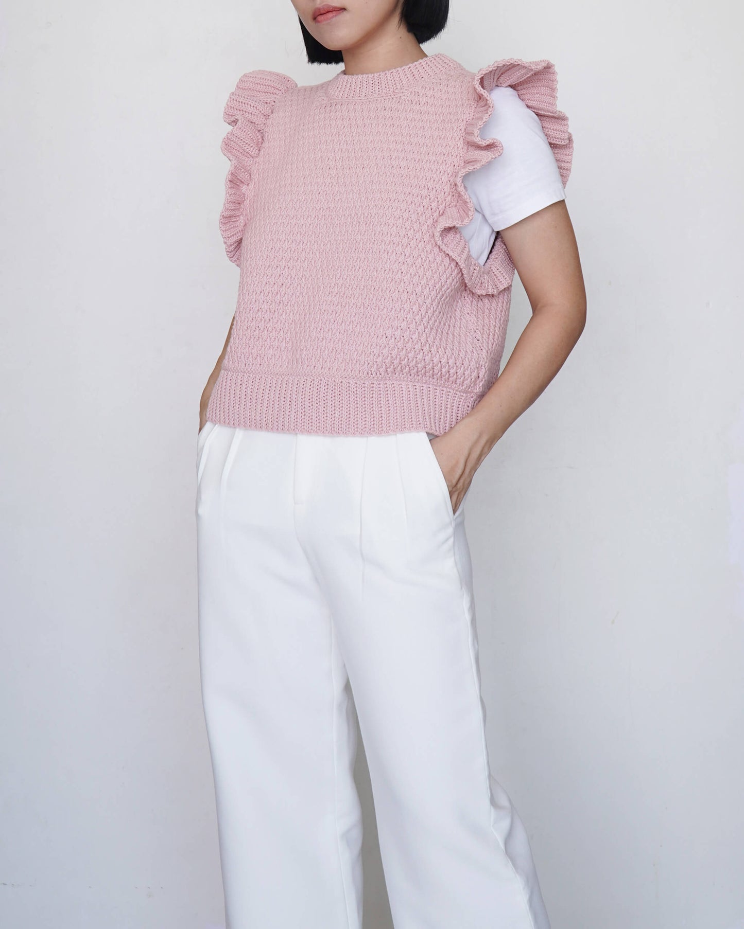 Person wearing a pink ruffled vest and white pants against a plain background